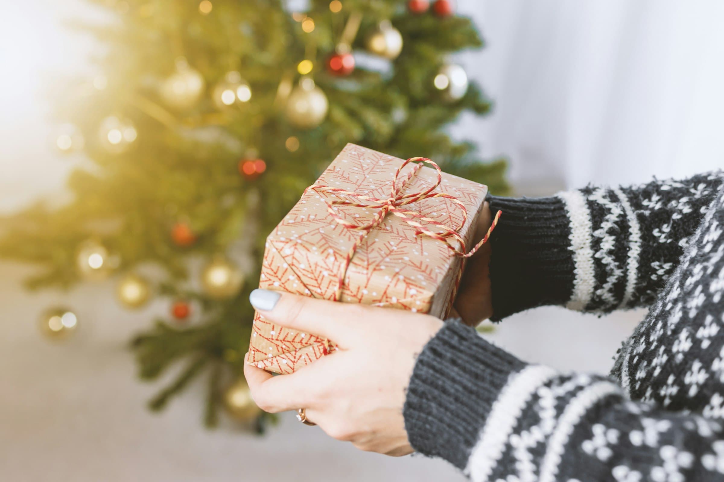 Manos con suéter navideño sostienen un regalo envuelto en papel kraft, frente a un árbol de Navidad